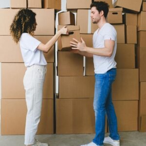 Side view of young lady with curly hair in casual clothes passing cardboard box to boyfriend during moving in new apartment in daylight