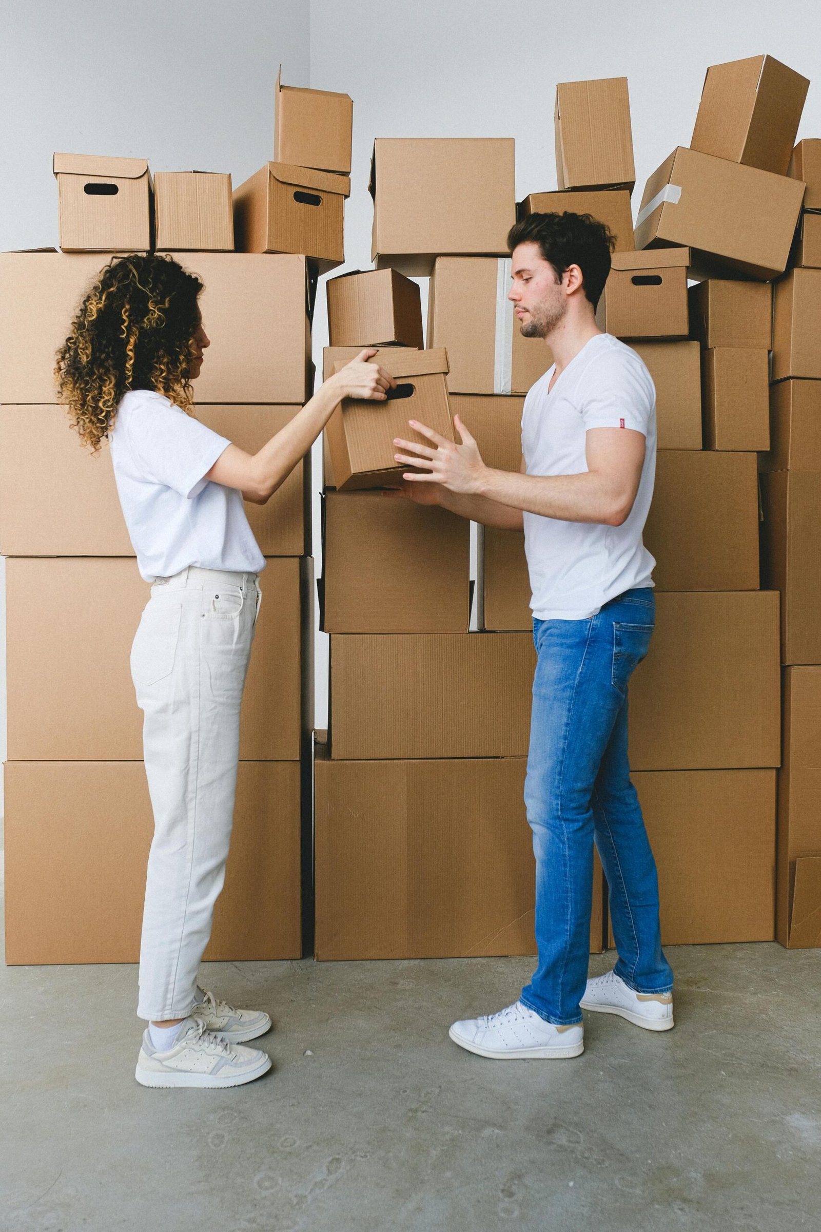 Side view of young lady with curly hair in casual clothes passing cardboard box to boyfriend during moving in new apartment in daylight